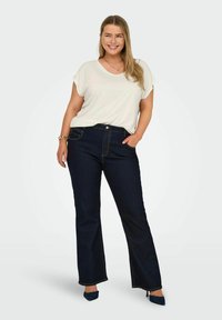 Un haut blanc à manches courtes associé à un jean évasé bleu foncé. Le jean a une taille mi-haute, des surpiqûres contrastées et un léger brillant.