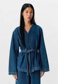 Veste en denim à manches longues, décolleté en V profond et ceinture nouée. Présente une silhouette en peplum et des coutures contrastantes. Couleur bleu profond.