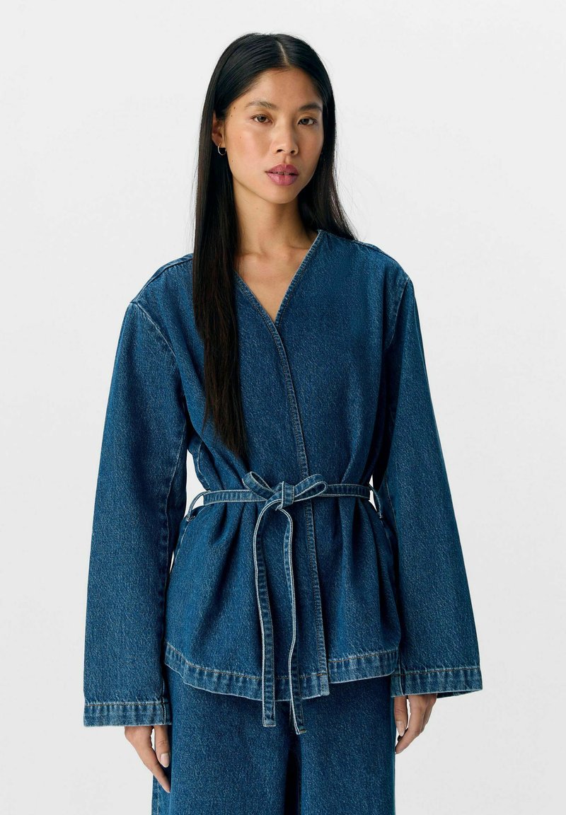 Veste en denim à manches longues, décolleté en V profond et ceinture nouée. Présente une silhouette en peplum et des coutures contrastantes. Couleur bleu profond.