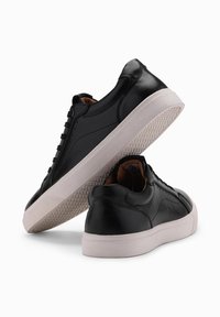 Baskets en cuir noir avec semelles en caoutchouc blanc, présentant des bouts ronds, des lacets plats et de subtiles perforations sur les côtés.