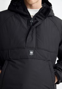 Giacca pullover nera con cappuccio, dotata di tasca frontale e etichetta con logo. Realizzata in tessuto liscio con una texture trapuntata e chiusura a zip.