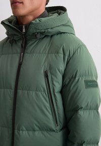 Zielona kurtka puchowa z kapturem, wyposażona w kieszeń na zamek, teksturowaną tkaninę oraz naszywkę z logo na rękawie.