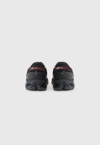 Sapatos atleticos pretos com um cabedal em malha, salto arredondado e sola preta brilhante. Apresenta marcas vermelhas no calcanhar e acabamentos texturizados.