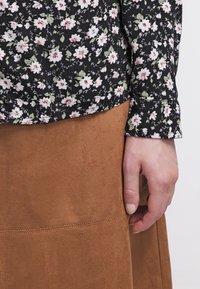 Main reposant à côté d'une jupe en suédine marron, portant un haut noir à manches longues avec un petit motif floral blanc et rose.