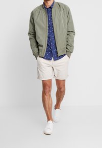 Zielona oliwkowa kurtka bomber, granatowa koszula w wzory, beżowe szorty na rolce i białe sneakersy. Luźny strój z różnorodnymi teksturami i kolorami.