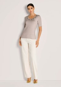 Vrouw staat tegen de muur in een taupe top met korte mouwen en juwelen op de hals, witte op maat gemaakte broek en metalen gouden hoge hakken.