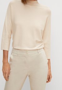Maglione lavorato a maglia beige chiaro con collo alto e maniche a tre quarti, abbinato a pantaloni beige su misura con chiusura a bottone.