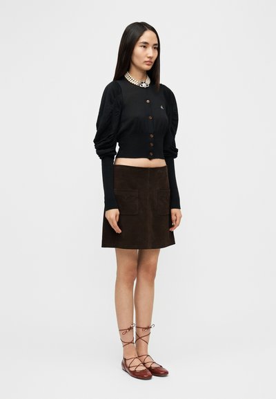 Μαύρο κοντό ζακέτα με φουσκωτά μανίκια και καφέ κουμπιά, συνδυασμένο με καφέ σουέντ φούστα με δύο τσέπες και κόκκινα δετά παπούτσια.