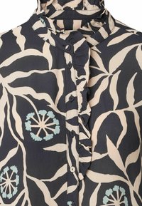 Blusa scura con motivo floreale e colletto a volants. Presenta bottoni lungo la parte anteriore e accenti verde pastello tra i colori beige e blu navy.