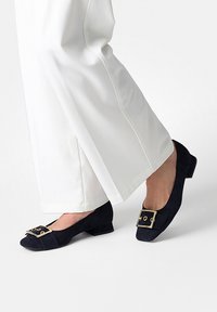 Chaussures bleu marine en daim à petit talon carré, ornées d'une boucle dorée et d'un détail plissé, associées à un pantalon large blanc.