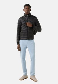 Sort vatteret dunjakke med høj krave, lyseblå bukser og beige sneakers. Modellen bærer en sort rygsæk.