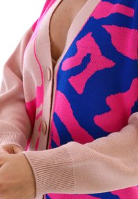 Stickad kofta med ett rosa och blått abstrakt mönster, mjuk textur, v-hals och två stora, ljusa knappar framtill.