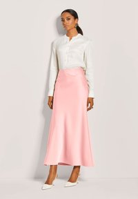 Zijde crèmekleurige blouse met knopen, gecombineerd met een satijnen, lichtroze maxi-rok. Witte pumps met puntige neuzen accentueren de outfit. Model staat recht vooruit gericht.