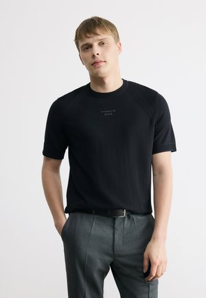 Fiatal férfi rövid szőke hajjal, fekete Porsche BOSS pólót és szürke nadrágot viselve, egyik kezét a zsebében tartva áll.