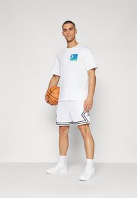 Bílý tričko s gradientním modrým grafikou, v kombinaci s bílými síťovanými basketbalovými šortkami s černými akcenty a bílými teniskami, držící basketbal.