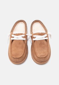 Paar pruune seemisnahast mokassiin-sussid valgete paelte ja pehme kreemja voodriga, valgel taustal.