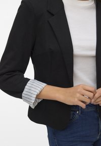 Schwarze Blazer mit tailliertem Schnitt, mit Reverskragen und gestreiftem Manschettendetail. Getragen über einem weißen Oberteil, kombiniert mit blauen Jeans.