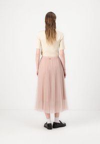Myk beige tettsittende topp og lang, plisserte rosa tyllskjørt. Antrekket er kombinert med svarte sko og hvite sokker, sett fra baksiden.