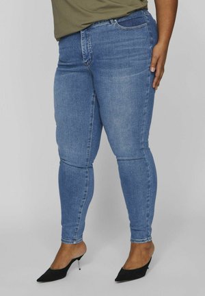 Jeans de mezclilla azul ajustados con una textura suave y un ligero desgaste. Presentan un diseño de cinco bolsillos y una cintura estándar. Combinados con tacones negros de punta.