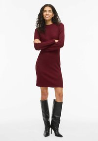 Abito maglione color bordeaux con maniche lunghe e scollatura rotonda, abbinato a stivali neri con tacco a spillo al ginocchio per una silhouette strutturata.