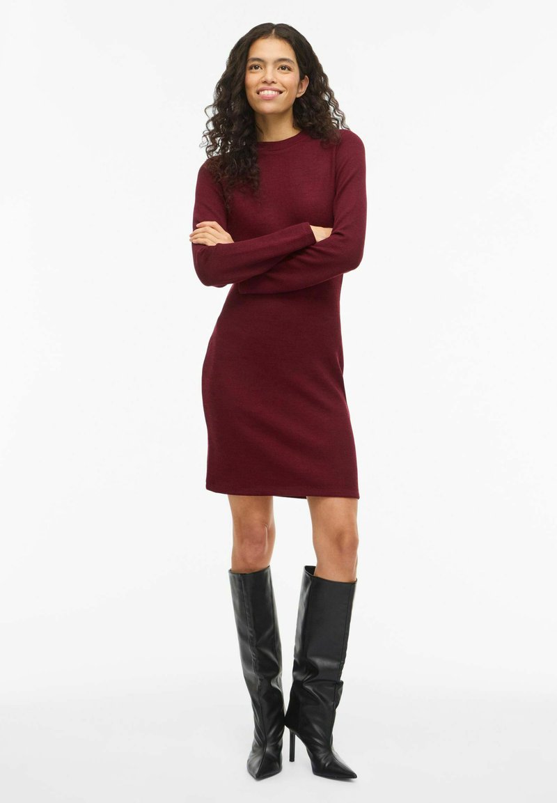 Abito maglione color bordeaux con maniche lunghe e scollatura rotonda, abbinato a stivali neri con tacco a spillo al ginocchio per una silhouette strutturata.