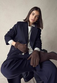 Brown suede gloves with stitching details, worn with a navy blazer over a white sweater and gray striped shirt, against a neutral background.