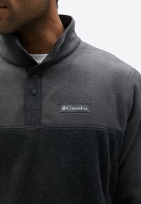 Bărbat purtând un pulover din fleece gri închis și negru, cu guler înalt și nasturi capse, cu un patch cu sigla Columbia pe piept.