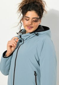 Světle modrá zipová bunda s vysokým límcem a nastavitelnou kapucí. Obsahuje černé detaily, přední zip a strukturovanou tkaninu.