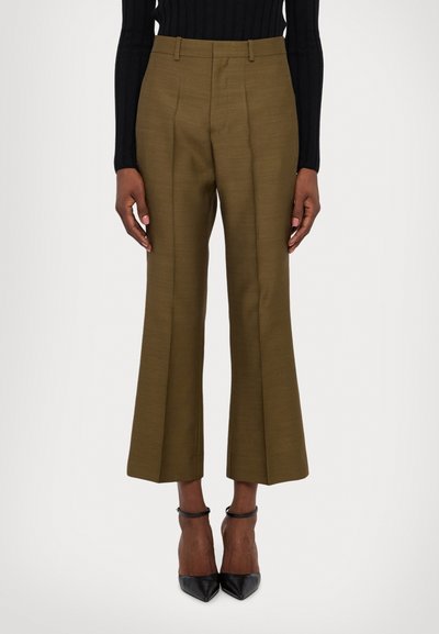 Pantaloni tailleur verde oliva con design a gamba svasata, caratterizzati da una texture liscia e pieghe visibili, abbinati a tacchi a punta neri.