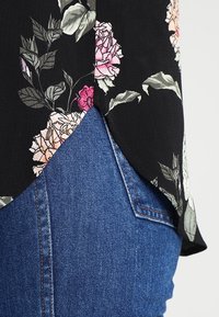 Blusa negra con estampado floral de rosas rosas y blancas, con un dobladillo curvado y una tela ligera y texturizada, llevada sobre unos jeans de denim azul.