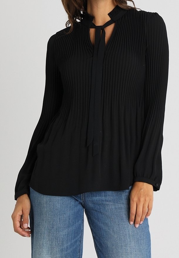 Blouse noire plissée à manches longues avec col volanté et nœud devant, portée avec un jean bleu.