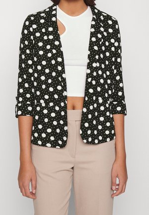 Zwarte blazer met witte stippen, lange mouwen opgerold bij de manchetten, getailleerde pasvorm, gecombineerd met een witte crop top en beige broek.