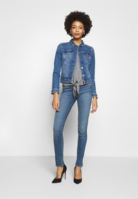 Woman with curly hair wearing a blue denim jacket, checkered blouse tied at waist, blue skinny jeans, and black high heels, standing against white background.