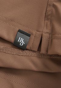 Brauner Stoff mit glatter Textur, der eine genähte Kante und ein schwarzes Etikett mit weißer Schrift zeigt. Der Stoff wirkt matt und strapazierfähig.