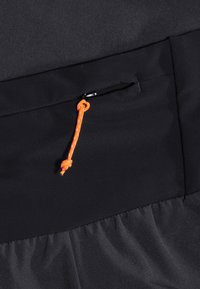 Svart tyg med en slät yta, med en dragkedjeficka som pryds av en orange paracorddragkedja. Synliga stygn längs sömmarna.