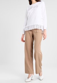 Vit långärmad topp med volangkant och logotyp av strass, kombinerad med beige byxor med vida ben och vita skor med klossklack.