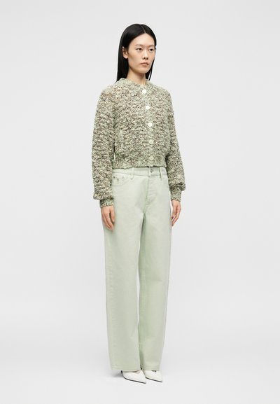 Femme debout, portant un cardigan vert texturé boutonné, un pantalon vert clair à jambes larges et des chaussures blanches à bouts pointus, sur fond uni.