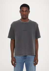 Grå bomulds T-shirt med rund hals, korte ærmer og sort logo på forsiden. Løs pasform med synlige syninger.