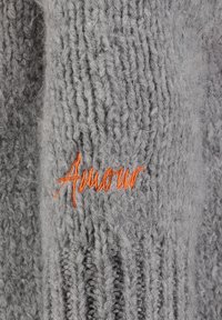 Close-up van grijs gebreid textiel met het oranje geborduurde woord "Amour" in cursieve letters.
