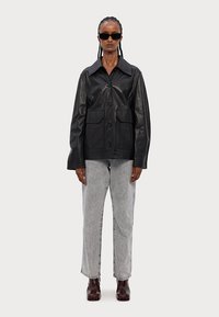 Must musträimiga musta nahkjaki, ees nööbiga kinnitamine ja kaks suurt taskut, koos heledate hallide teksade ja tumedate kingadega.