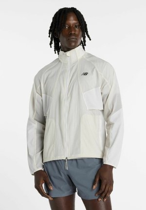 Person trägt eine weiße Zip-up-Jacke mit New Balance-Logo und graue Sportshorts, steht vor einem einfarbigen Hintergrund.