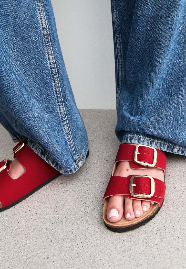Sandali di tessuto rossi con due fibbie, abbinati a jeans in denim blu. I sandali presentano una suola piatta e un design aperto sulla punta.