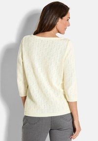 Cremefarbener Strickpullover mit 3/4 Ärmeln, ausgestattet mit einem strukturierten Spitzenmuster und ribbelförmigem Saum. Kombiniert mit grauen Hosen, von hinten betrachtet.