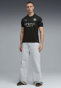 Jovem está de pé, vestindo uma camisa de futebol preta do Manchester City, calças jeans largas azul claro e sapatilhas pretas, contra um fundo cinza liso.