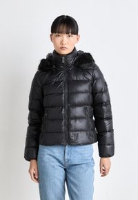 Giubbotto piumino nero con finitura lucida, dotato di cappuccio con pelliccia, chiusura con zip e tasche laterali, indossato sopra jeans blu.
