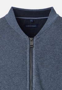Graue Strickjacke mit Rei�ßverschluss und geripptem Kragen. Sie hat eine gewebte Textur und ein kontrastierendes marineblaues Futter mit einem Etikett, auf dem "CASA MODA" steht.