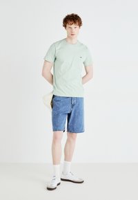 Camiseta de algodón verde menta con un pequeño logotipo, combinada con pantalones cortos de mezclilla azul. Presenta calcetines blancos y zapatillas blancas con acentos marrones.