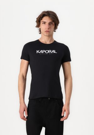 Jonge man met halflang haar, gekleed in een nauwsluitend zwart T-shirt met de tekst "KAPORAL summer sun", staand tegen een effen witte achtergrond.