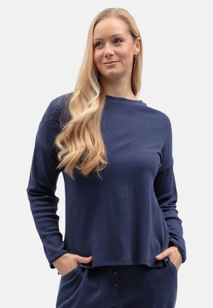 Blonde vrouw met lang golvend haar draagt een marineblauwe top met lange mouwen en bijpassende broek met handen in de zakken, kijkt iets naar rechts.