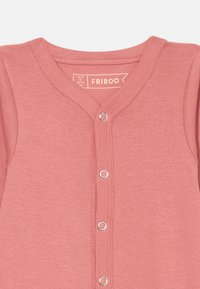 Růžový bavlněný overal s výstřihem do V, zapínáním na patentky a žebrovanou texturou. V oblasti výstřihu má štítek "FRIBOO".